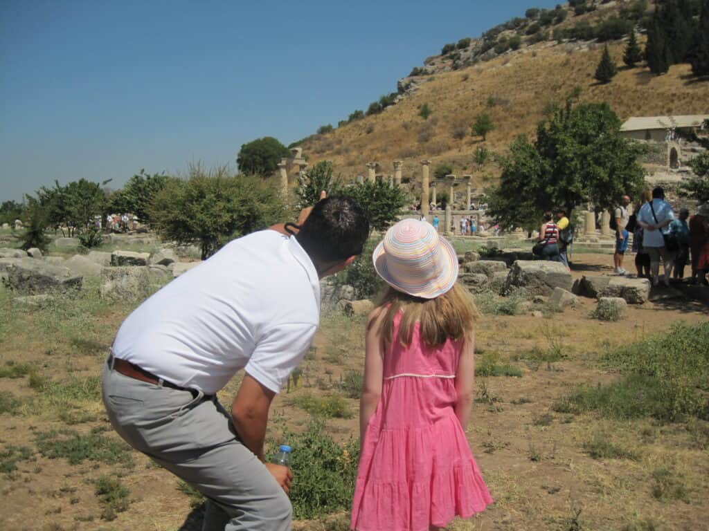 Young girl in dark pink dress and straw hat stands beside man in white t-shirt and gray trousers crouched down and pointing to ruins at Ephesus. 