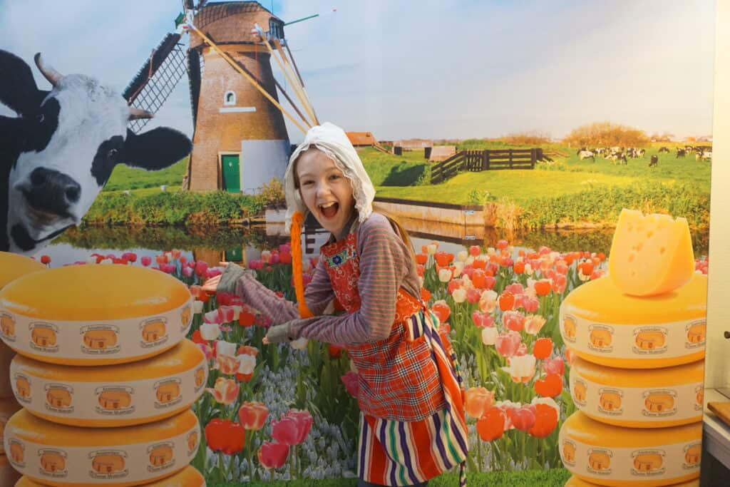 Young girl dressed in Dutch farm girl costume of apron and bonnet standing in front of photo of cheese, cows, tulips and windmill.