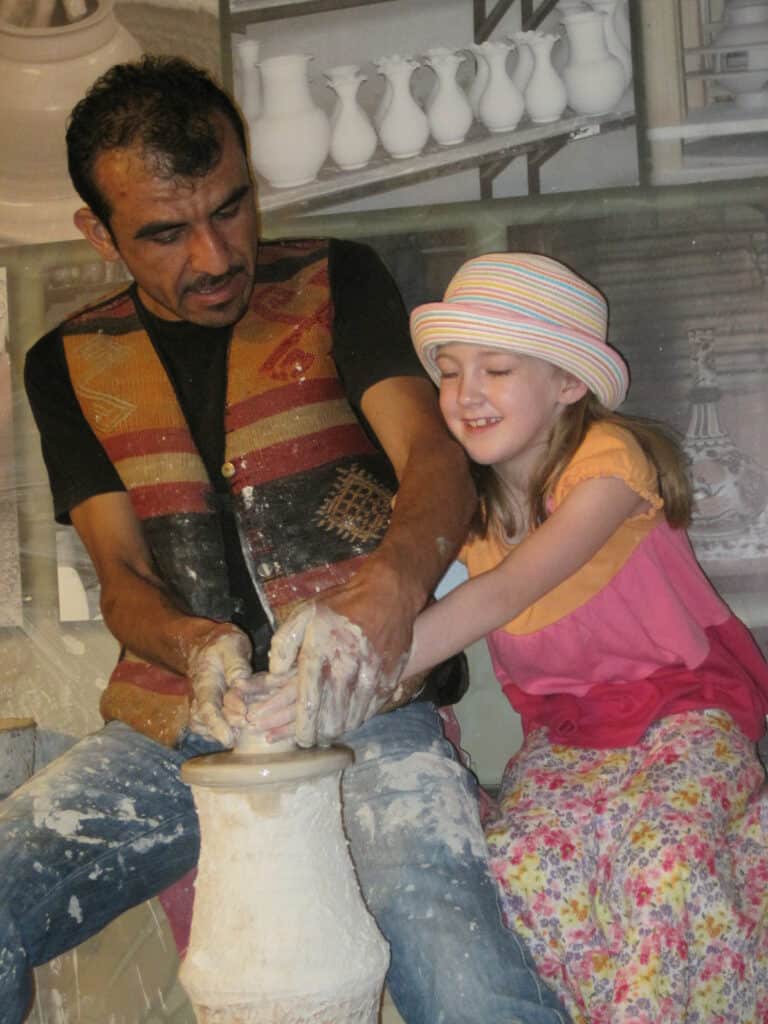 Man in black shiirt and vest helps young girl wearing multi-coloured hat, flowered skirt and pink and orange top sculpt a vase on a pottery wheel.