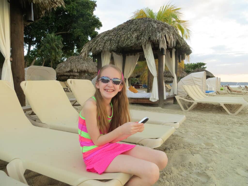 Smiling young girl wearing pink and green striped skirt and top and sunglasses sitting on beige coloured beach chair at Beaches Negril, Jamaica.