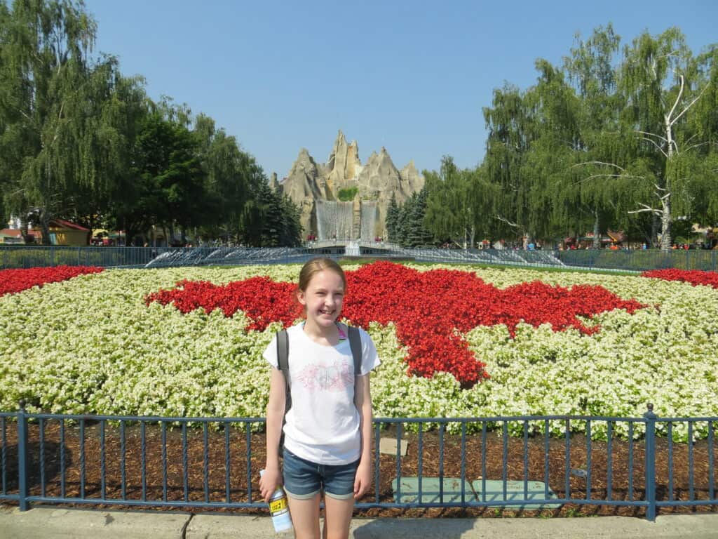 Young girl in denim shirts and t-shirt holding water bottle stands in front of garden designed to look like Canadian flag at Canada's Wonderland with mountain in background.