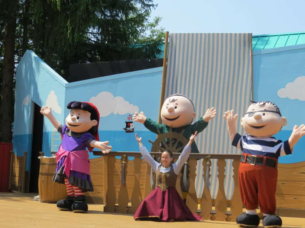 Female performer in shirt and vest kneeling on ground with arms upraised surrounded by three characters from Charlie Brown in pirate costumes on stage at Canada's Wonderland.