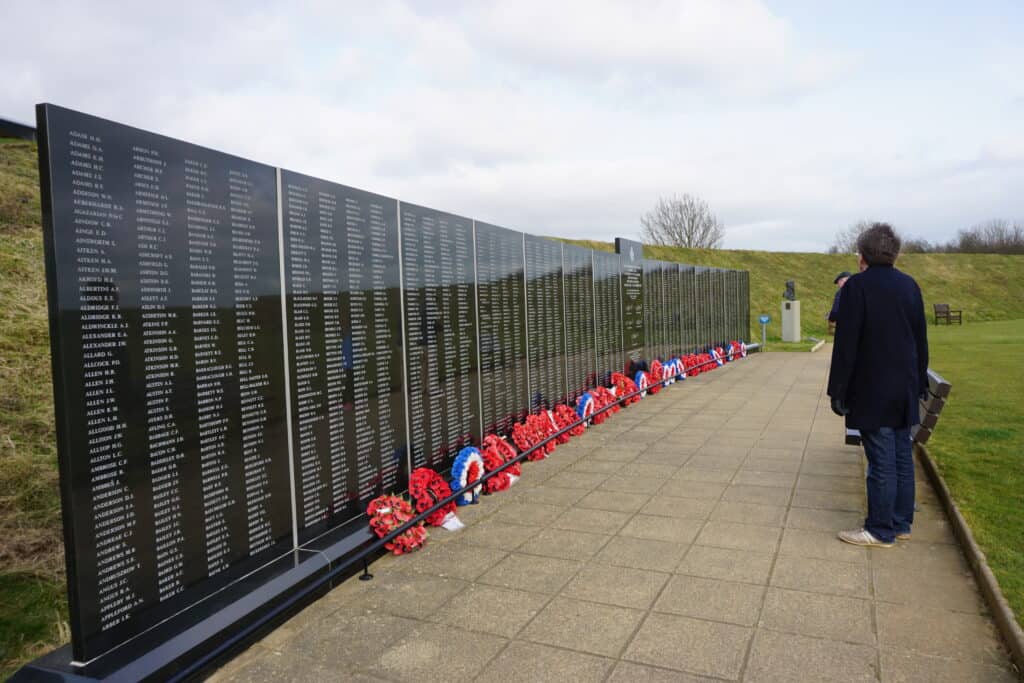 Man in dark clothes standing in front of Battle of Britain Memorial Wall which has names etched on black wall and wreaths of poppies on ground along length of wall.