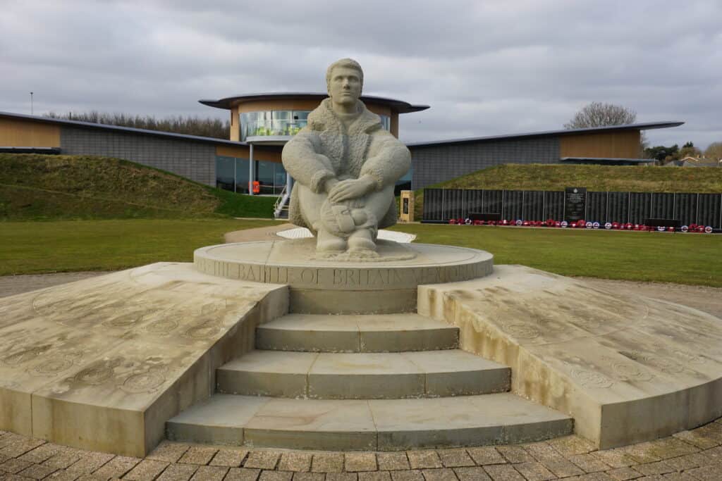 Sculpture of male pilot sitting on platform with words The Battle of Britain Memorial inscribed and building and Memorial Wall in background.