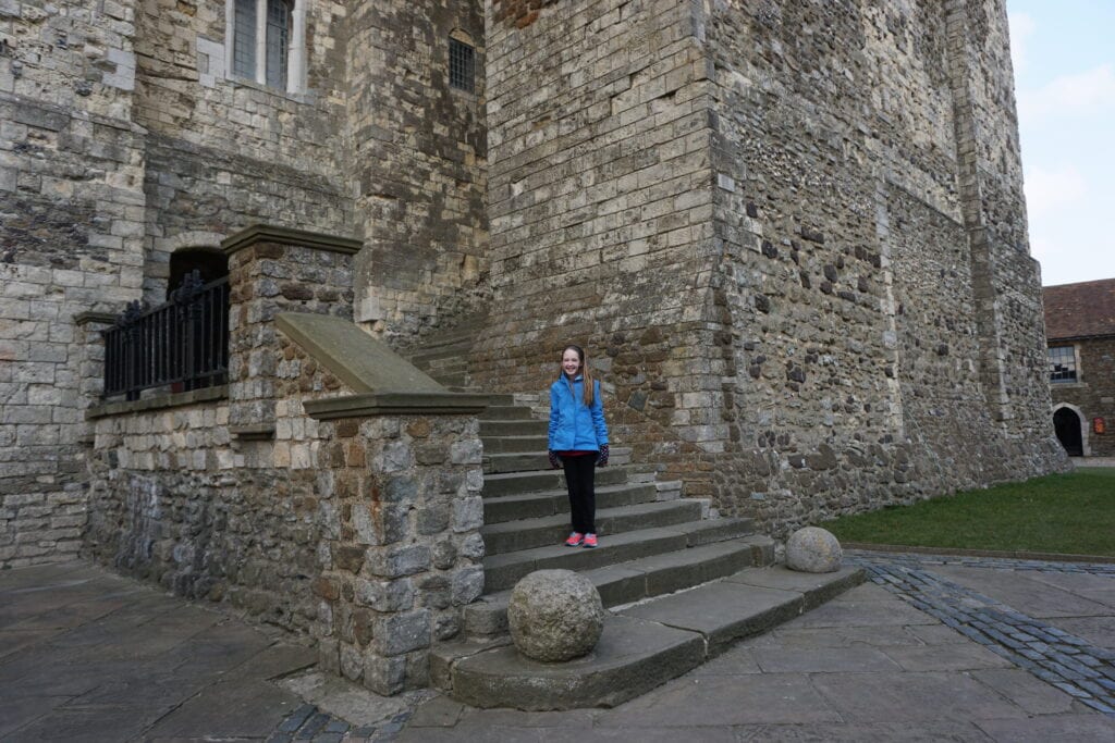 Young girl in blue coat and black pants standing on steps of stone building at Dover Castle, England.