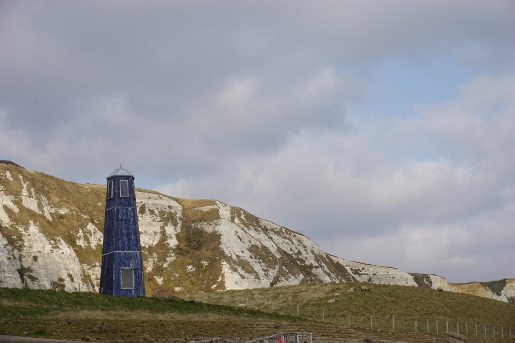 Blue coloured tower on hills in front of white cliffs partially covered with grass and sheeps grazing in distance.