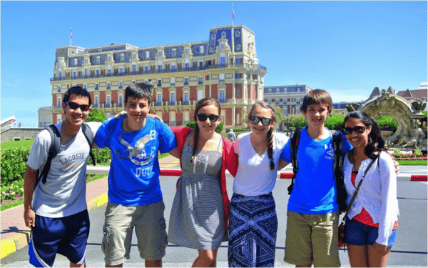 Group of 6 teens posing with buildings in background.