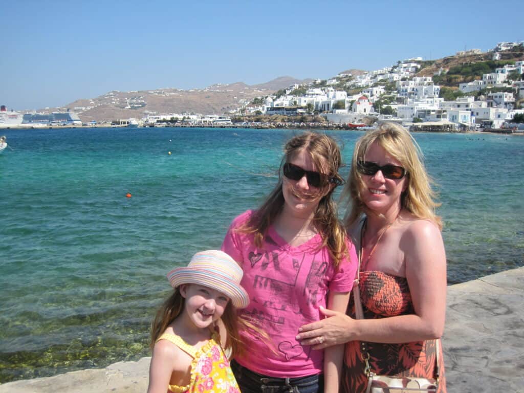 Woman wearing dress and sunglasses, teen girl in dark pink t-shirt and sunglasses, young girl wearing striped straw hat standing on waterfront in Mykonos town with sea and white buildings in background.