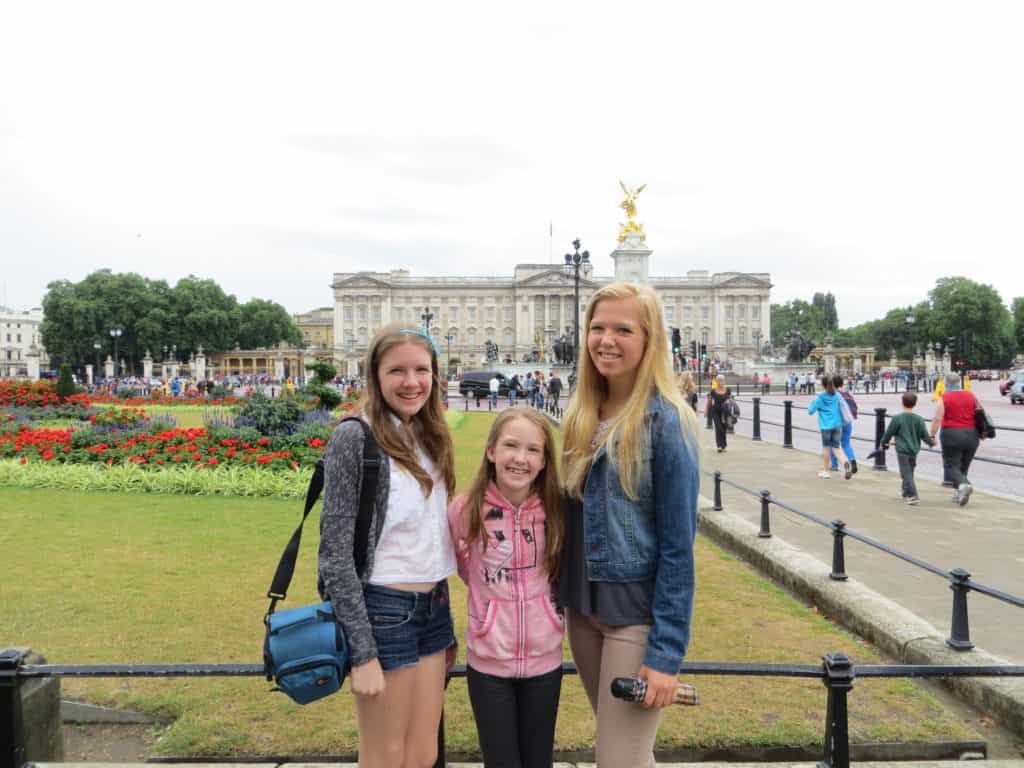 Two teen girls and a younger girl standing with flower gardens, crowds of people and Buckingham Palace in background.