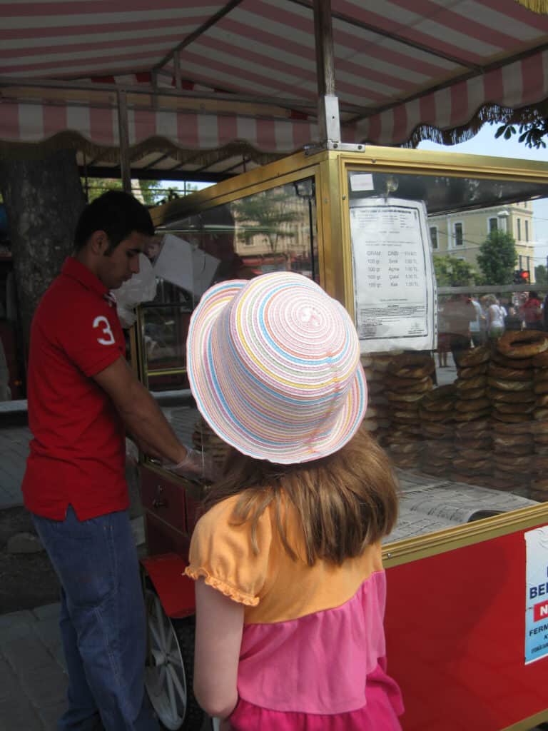 Young girl wearing multi-coloured striped straw hat buying a simit from a man in red shirt at a food cart in Istanbul, Turkey.