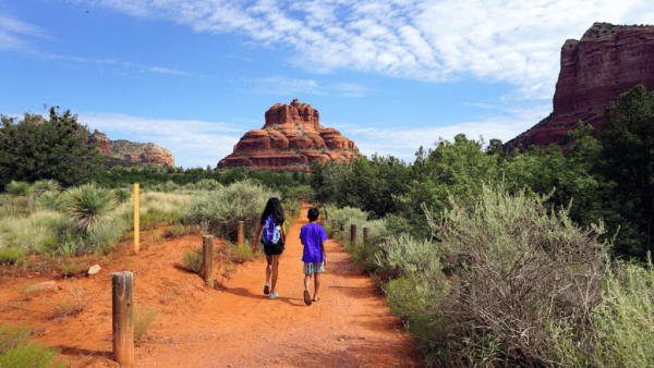 Two young kids walking on red dirt path through green vegetation toward hills in Sedona, Arizona.