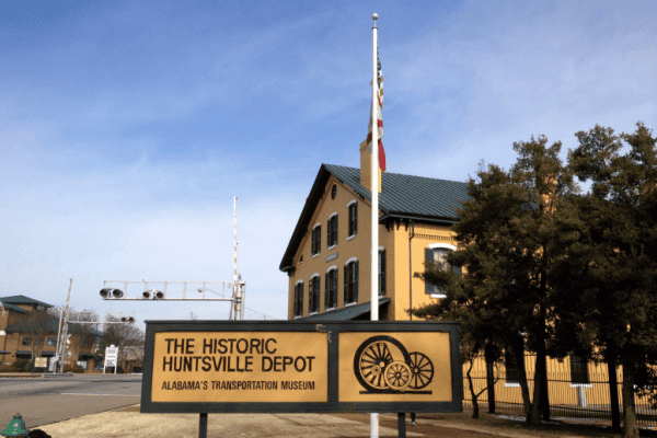 Dark yellow and green trimmed sign reading The Historic Huntsville Depot - Alabama's Transportation Museum beside trees and in front of flag pole and building painted in same colours.