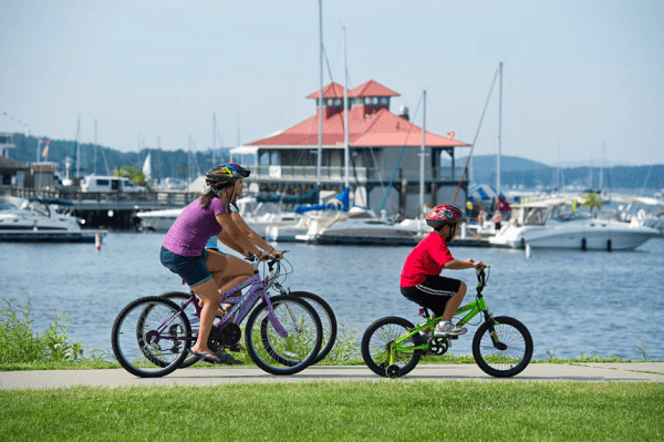 Two adults and a young boy cycling on bike path along waterfront in Burlington, Vermont with boats and building in background.