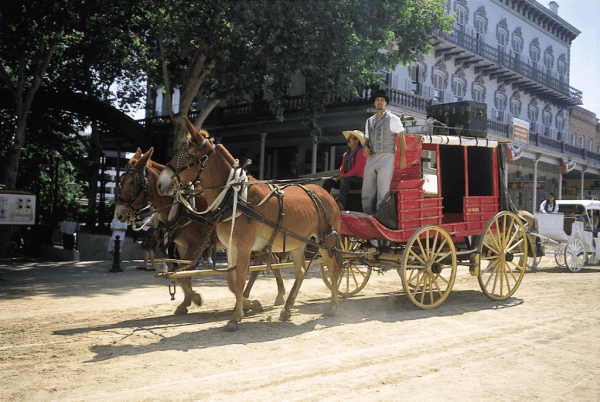 Two brown horses pulling a red carriage with hold coloured wheels with driver sitting and man standing in Old Sacramento.