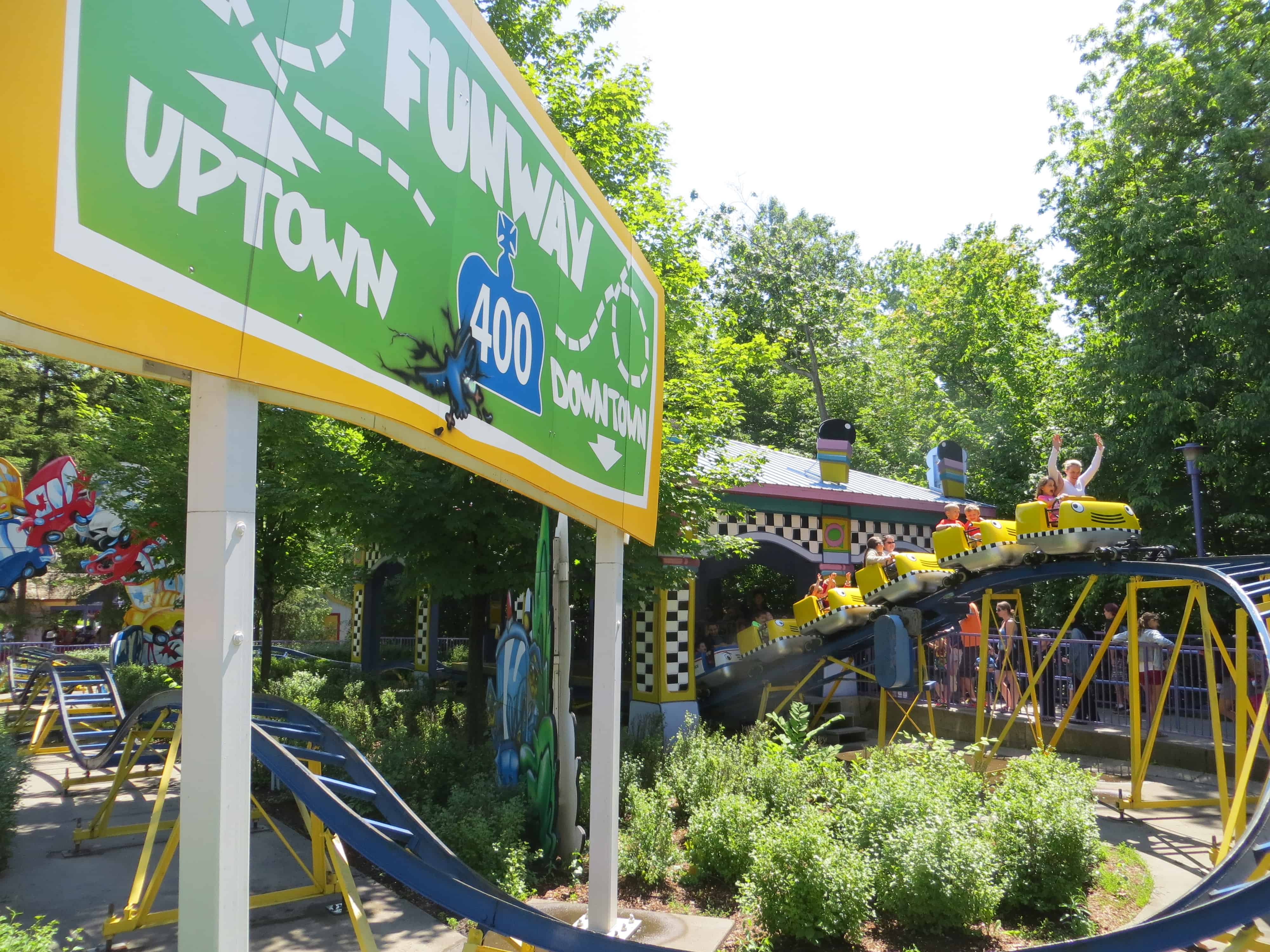 kids roller coaster at Canada's Wonderland - riders in yellow and black taxi cars coming around loop of ride with green and yellow highway sign in foreground.