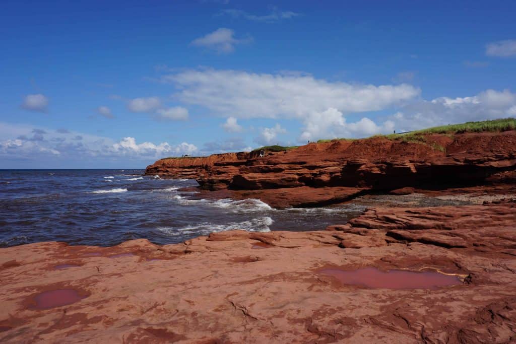 Red stone cliffs along ocean in Prince Edward Island with bright blue sky and a few clouds.