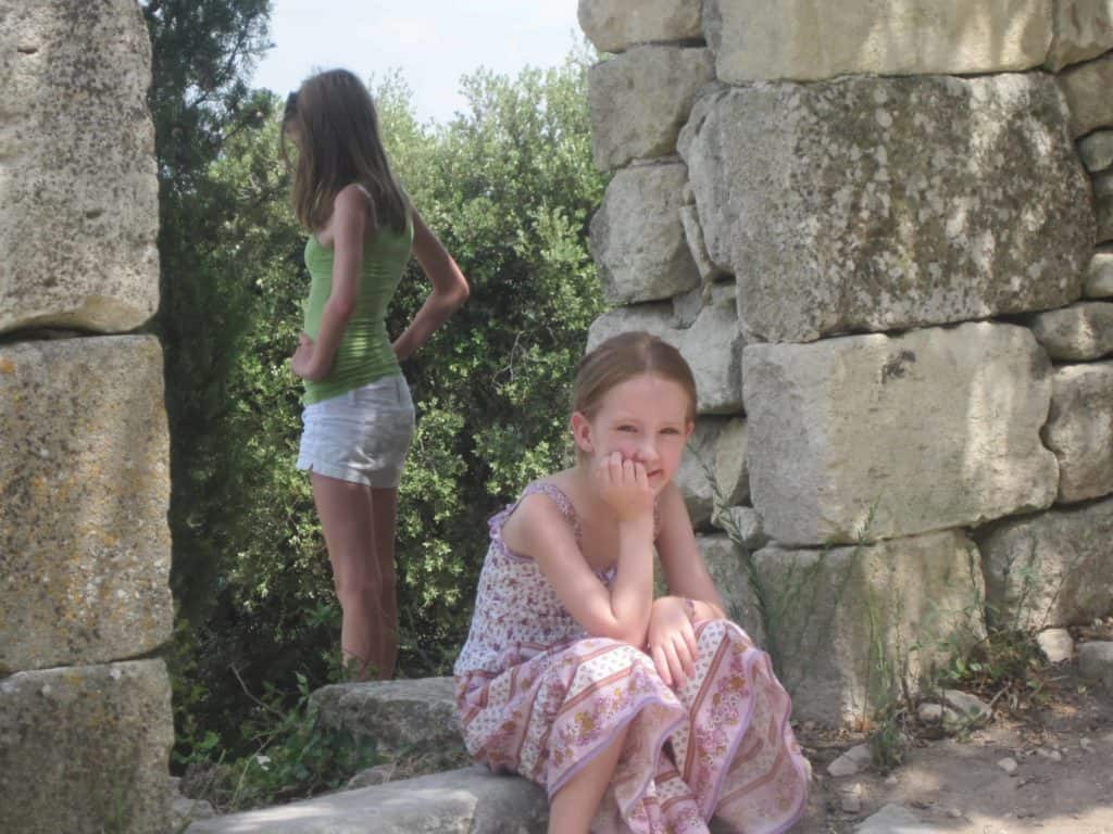 Young girl wearing purple flowered dress with hand on chin sitting in doorway of stone ruins with teenage girl in white shorts and green tank faced away in background.