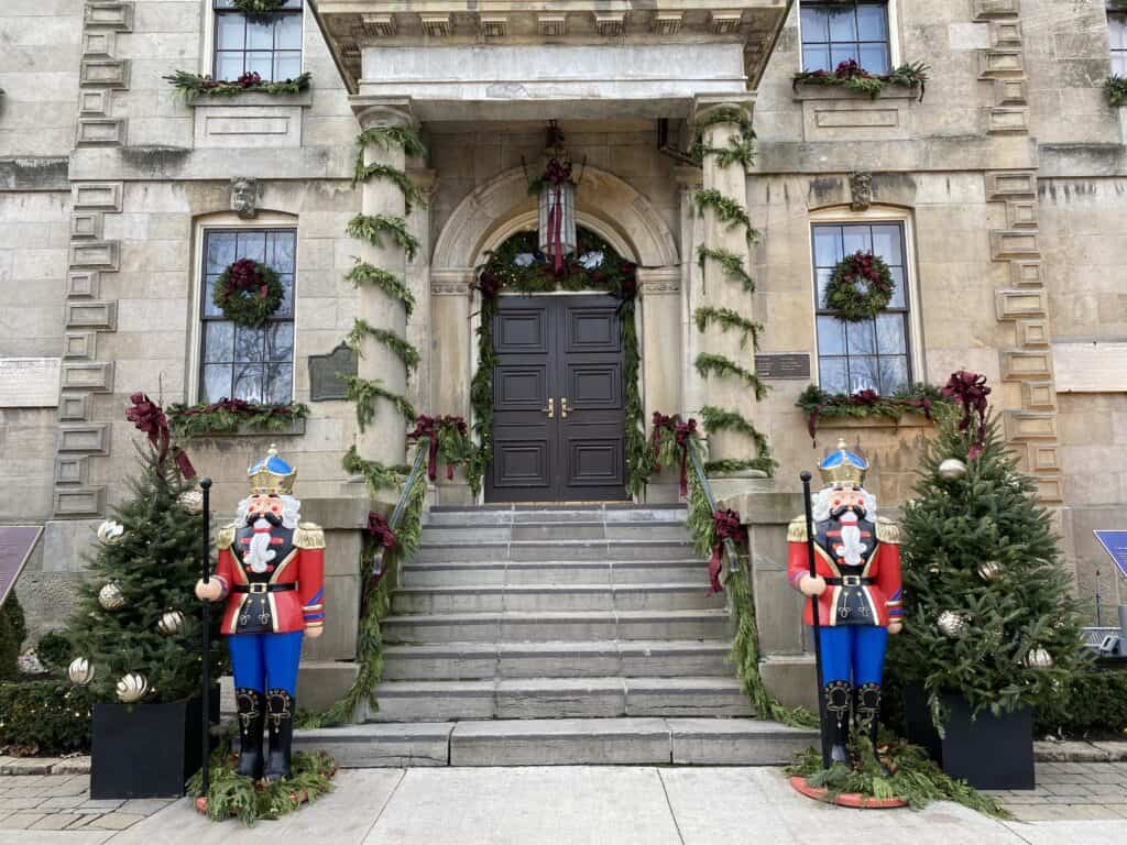 Facade of historic Court House in Niagara-on-the-Lake decorated for the holiday season - wreaths on windows greenery and burgundy bows and two Christmas trees and red and blue soldier statues flanking stairs to front entrance.