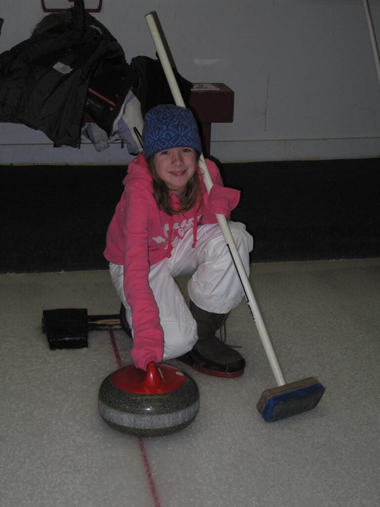 Young girl wearing toque in white snow pants and pink sweatshirt holding rock and broom on curling rink at chateau montebello.