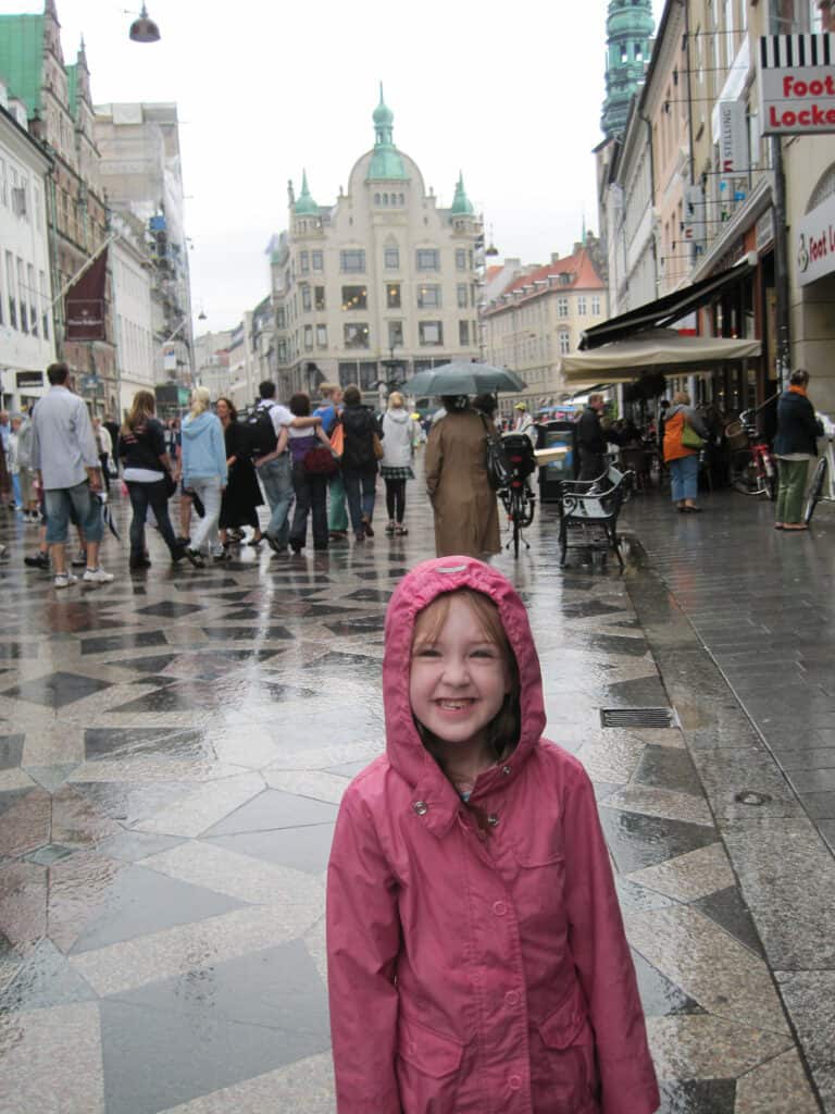 Young girl in pink raincoat with hood grinning while standing in rain in Copenhagen pedestrian shopping district.