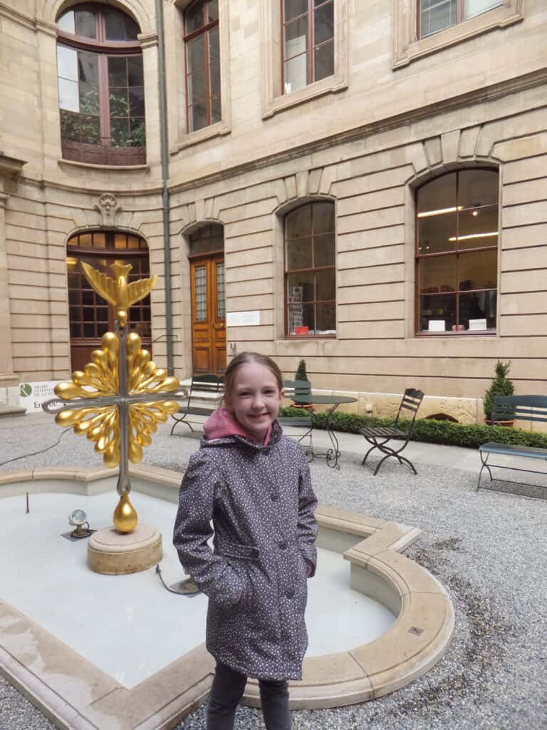 Young girl in grey flowered coat standing beside sculpture of golden dove in the courtyard of Maison Mallet in Geneva.