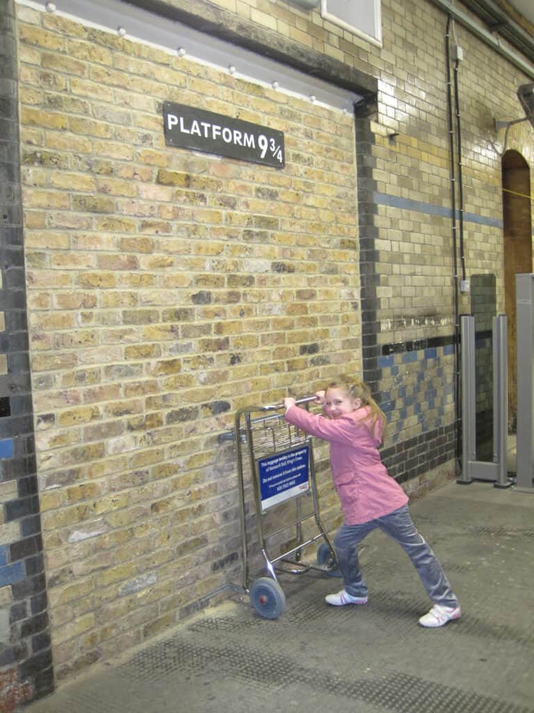Young girl in pink coat pushing luggage cart against brick wall beneath sign Platform 9 3/4.
