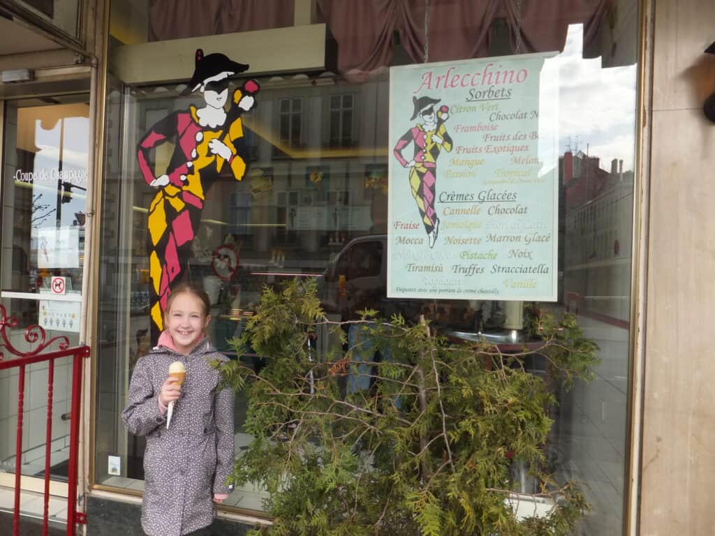 Young girl wearing grey flowered coat holding cone of gelato standing outside the Gelateria Arlecchino in Geneva, Switzerland.