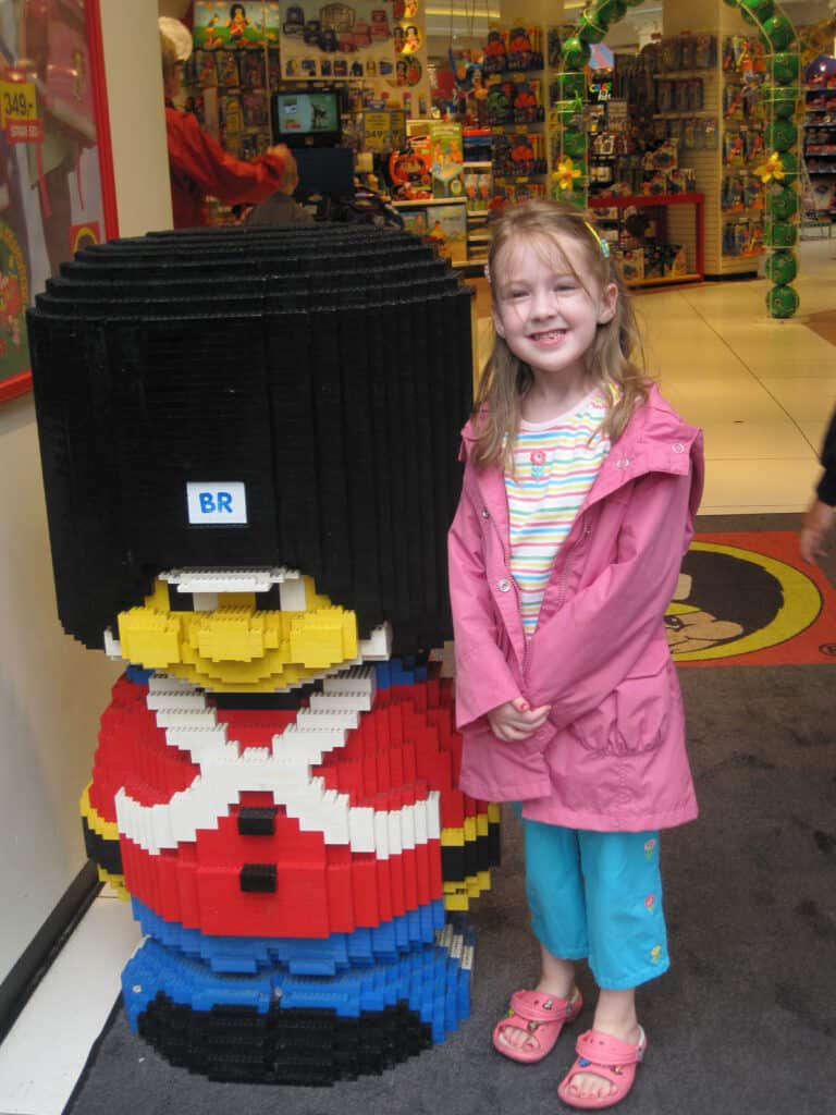 Young girl in pink coat and striped t-shirt stands beside LEGO guard outside LEGO shop in Copenhagen.