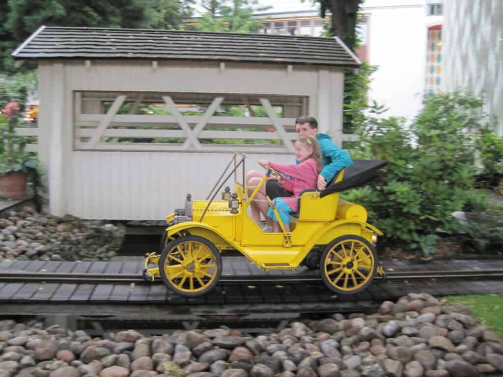 Dad sitting with young girl in pink coat driving bright yellow old-fashioned car on a track at Tivoli Gardens.