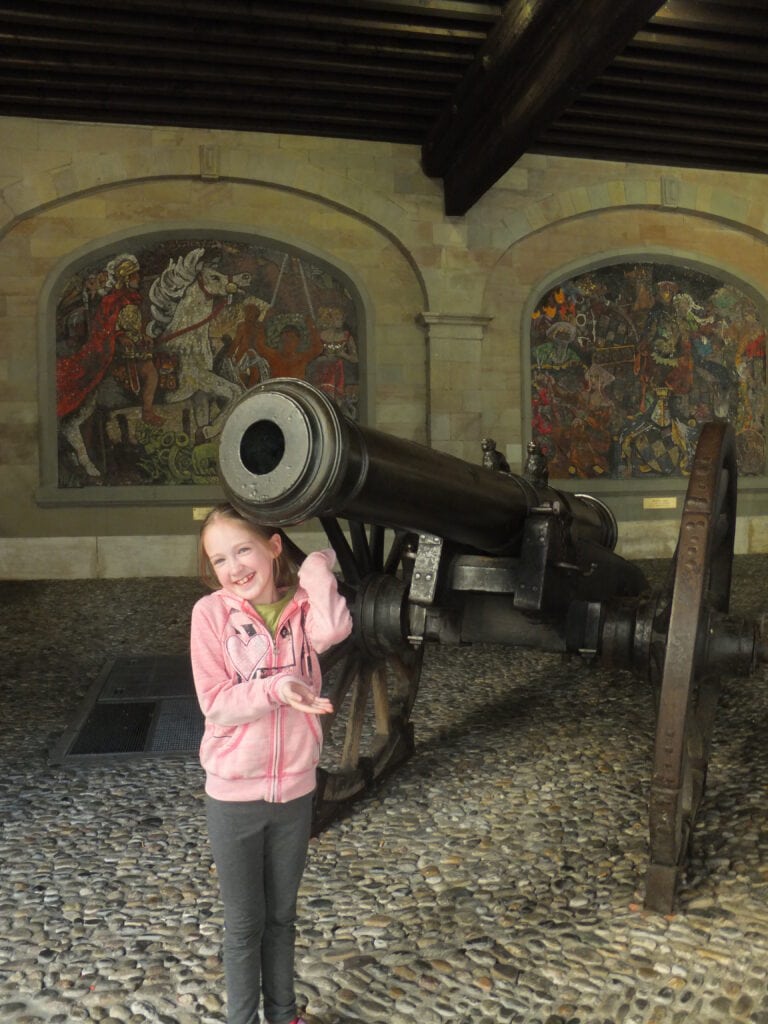 Young girl in grey pants and pink zipped sweatshirt poses in front of cannon at the Old Arsenal in Geneva, Switzerland.