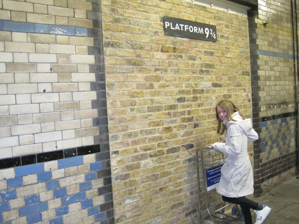 Young girl pushing luggage cart against brick wall beneath sign reading Platform 9 3/4 at Kings Cross station, London, England.