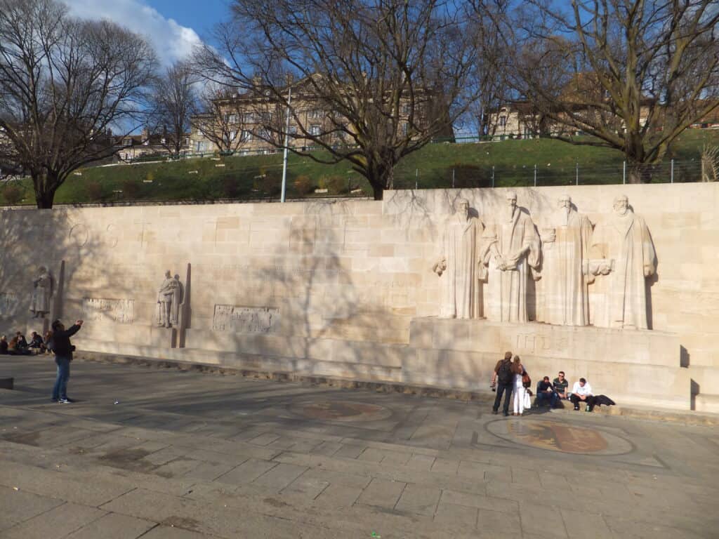 A few people standing and sitting in front of the Reformation Wall in Bastions Park in Geneva.