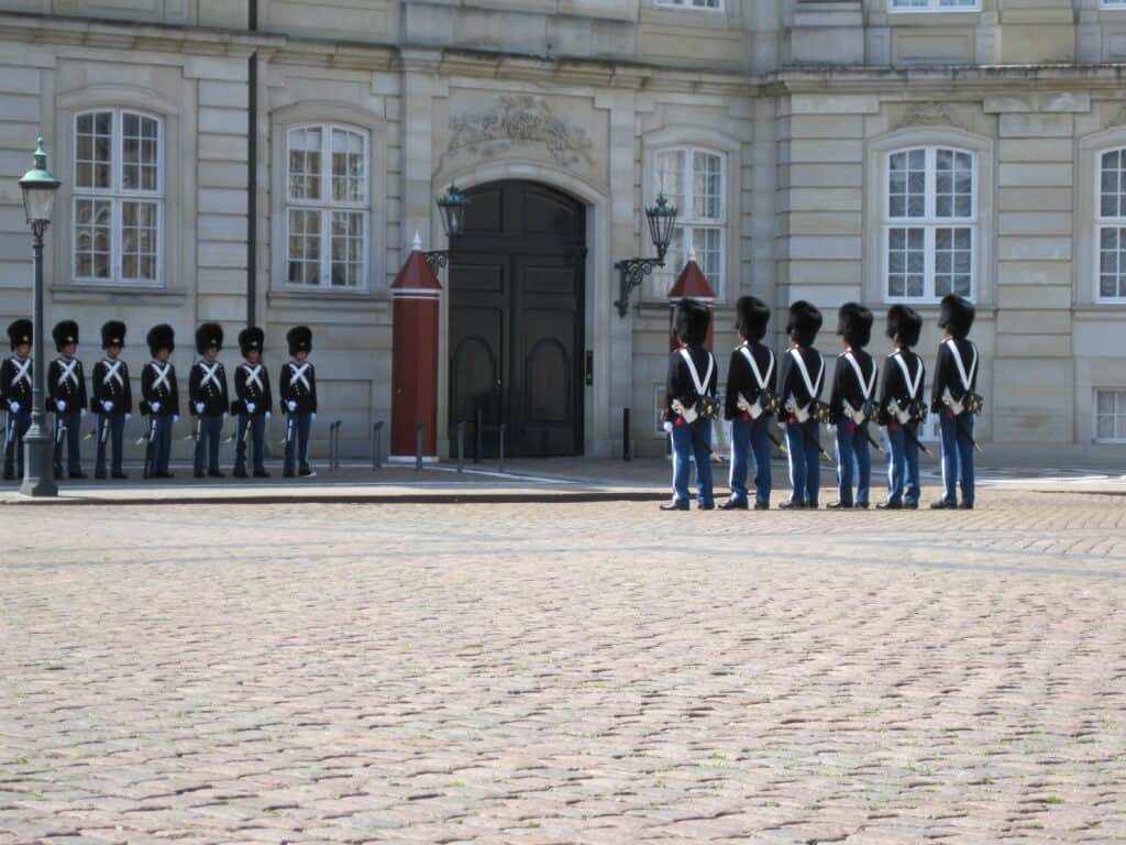 Two groups of guards wearing blue uniforms and black hats face each other in front of Amalienborg Palace in Copenhagen, Denmark.