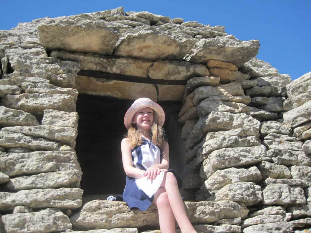 Young girl in blue and white dress and hat sits with legs crossed in window opening of a stone borie in Gordes, France.