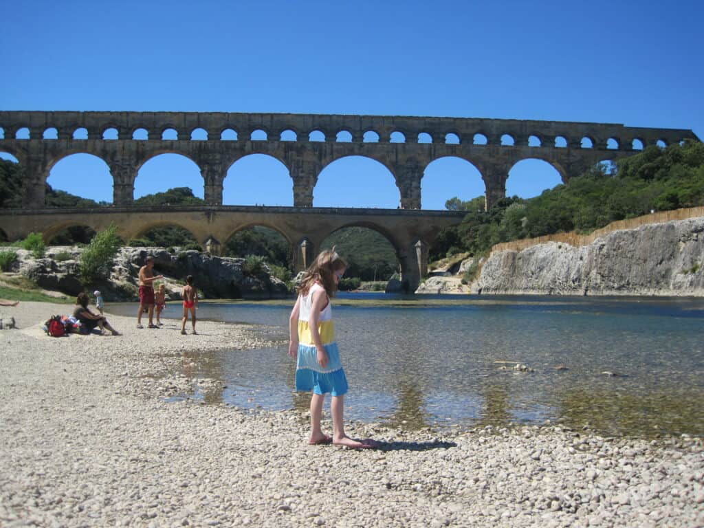 Young girl in blue, yellow and white striped dress standing at edge of water and a family in background at the Pont du Gard, France.