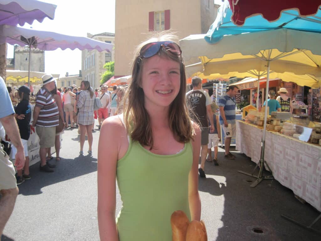 Teen girl in green tank top holding baguettes at market in Gordes, France.
