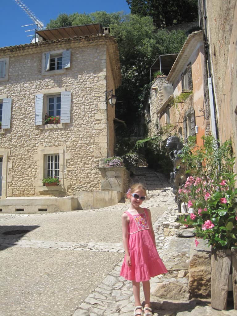Young girl in bright pink dress and sunglasses standing on cobblestone street beside bush with pink flowers and stone buildings in background - Joucas, France.