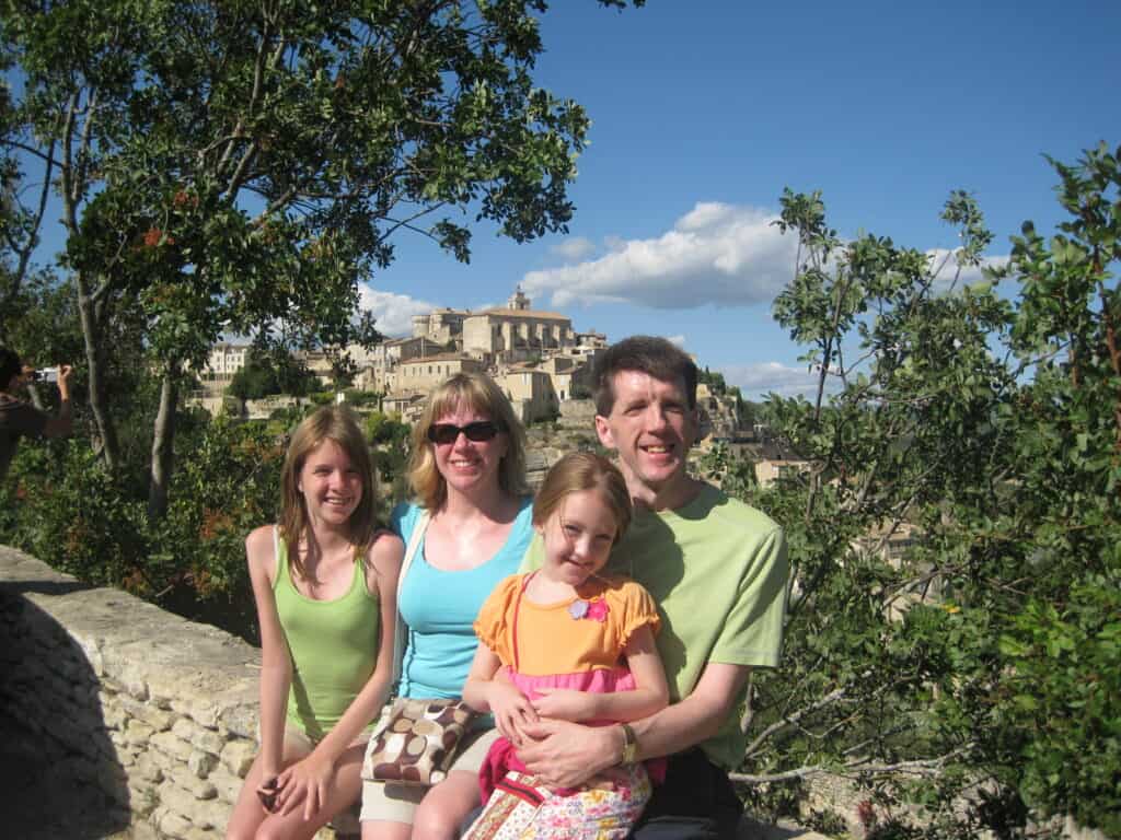 Family of mom, dad, teen girl and young girl sitting on stone wall with village of Gordes, France in background.