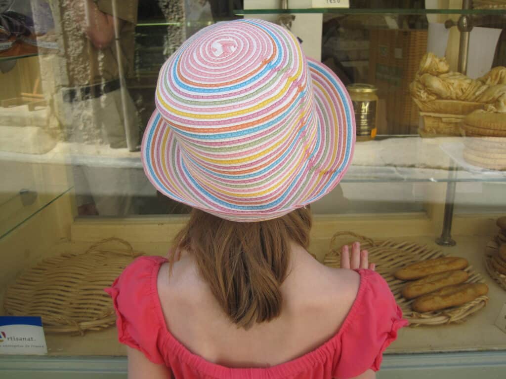 Young girl with coral top and multi-coloured striped straw hat looking in window of a bakery in Provence.