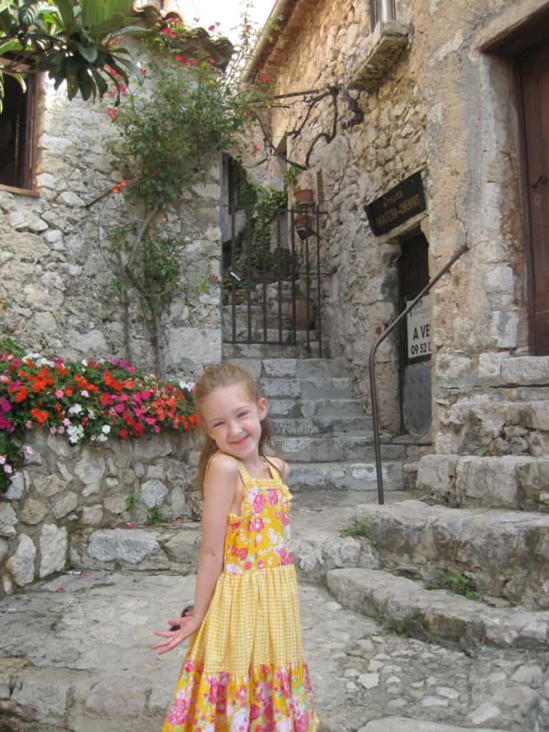Young girl in pink and yellow flowered dress standing in front of stone steps and planter of flowers in Eze, France.