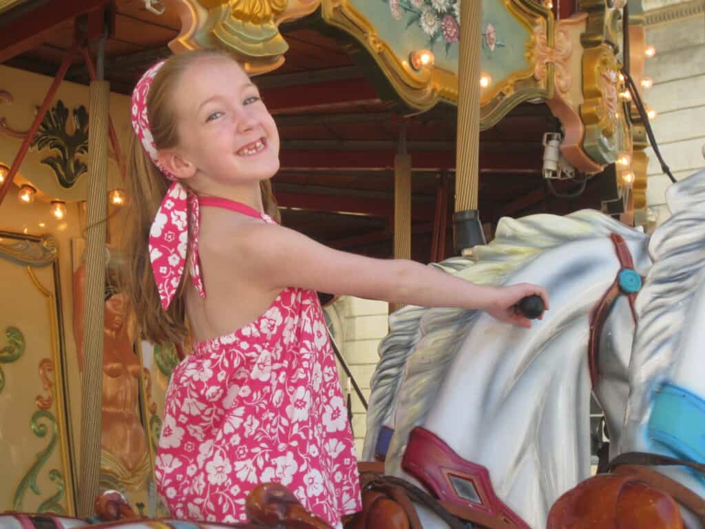 Girl in pink and white flowered top and matching headband sitting on a horse on the carousel in Avignon, France.