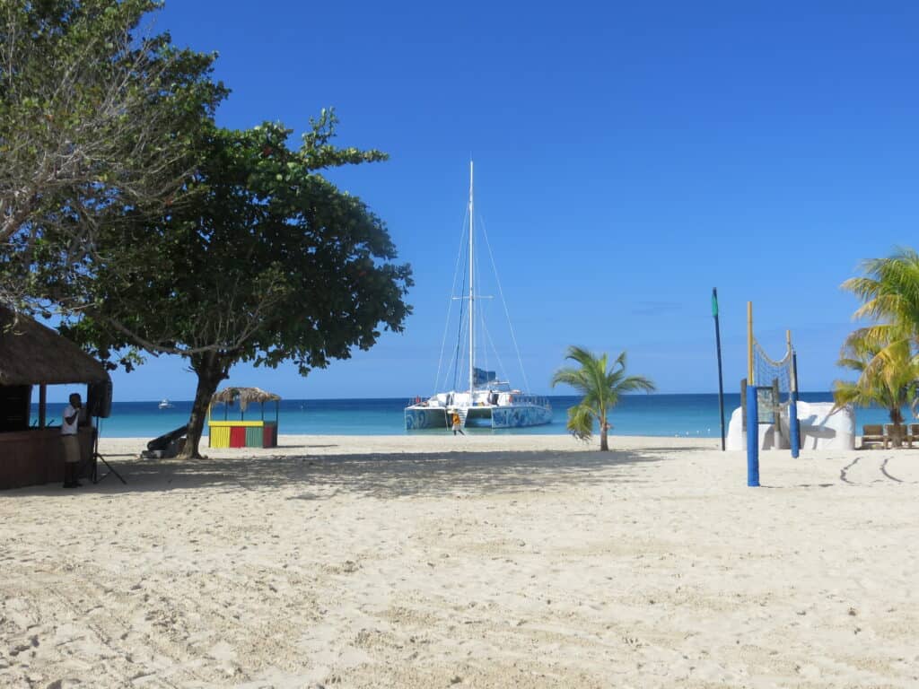 Quiet morning on beach at Beaches Negril - boat in water, volleyball net on sandy beach and a few trees with bright blue sky.