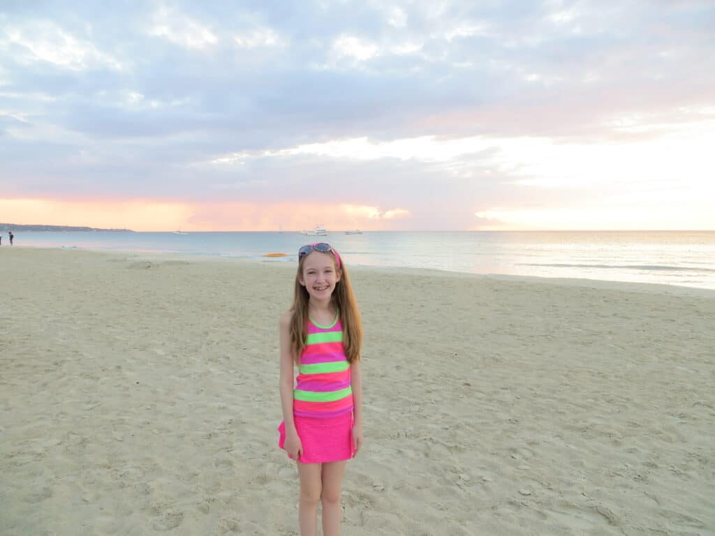 Young girl wearing bright pink skirt and pink and green striped top with sunglasses pushed up on head standing on white sand beach as sun begins to set at Beaches Negril resort.