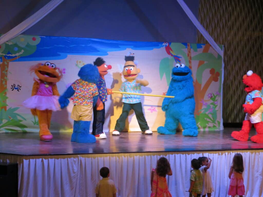 Small children stand in front of stage where group of Sesame Street characters including Elmo, Zoe, Cookie Monster, Bert and Ernie are performing.