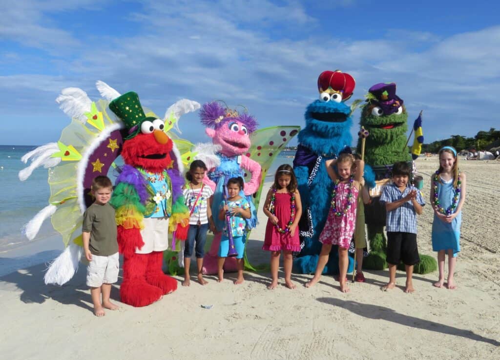 Group of children posing with four Sesame Street characters including Oscar, Cookie Monster and Elmo in Carnival costumes at Beaches Negril, Jamaica.