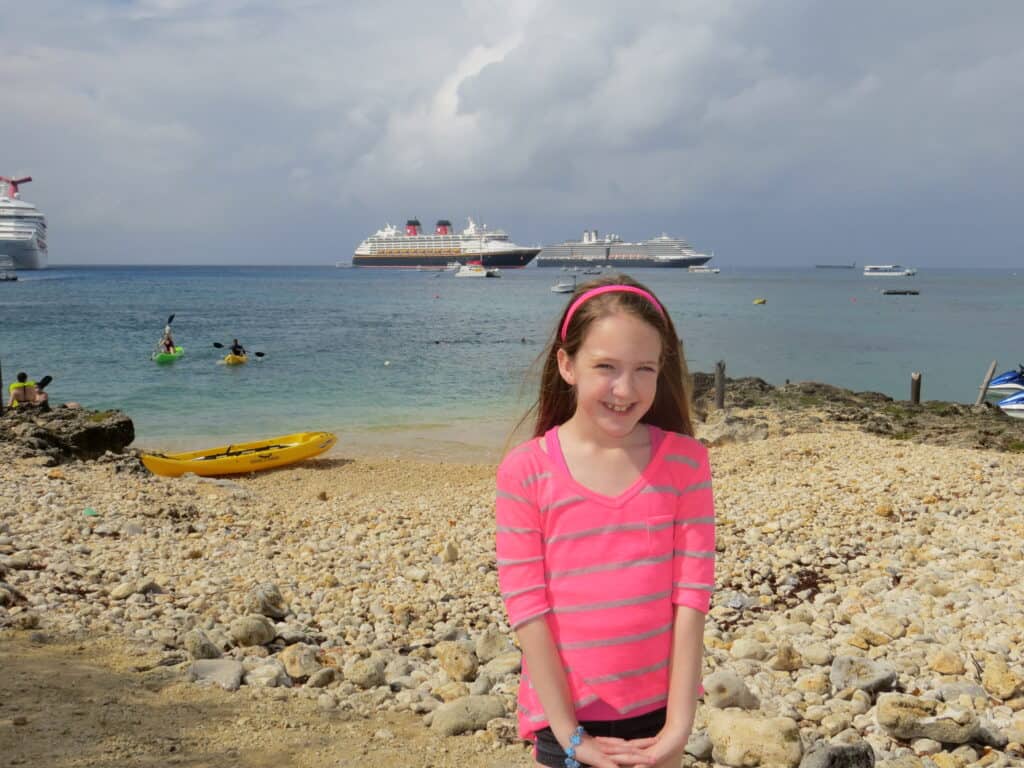 Young girl wearing pink and grey striped shirt and pink headband standing on shore in Grand Cayman with cruiseships in background.