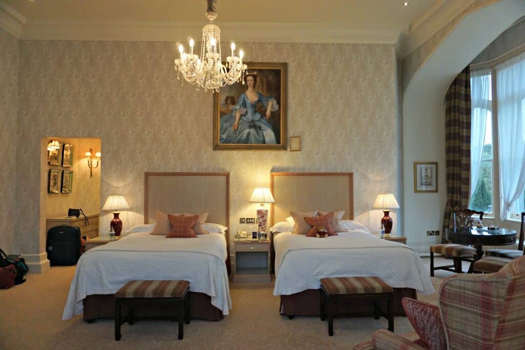 Room at Dromoland Castle with patterned cream wallpaper, glass chandelier over two beds with white linens and cream and pink cushions. Striped footstools at ends of bed, three night tables with lamps and painting of woman in blue gown over beds.