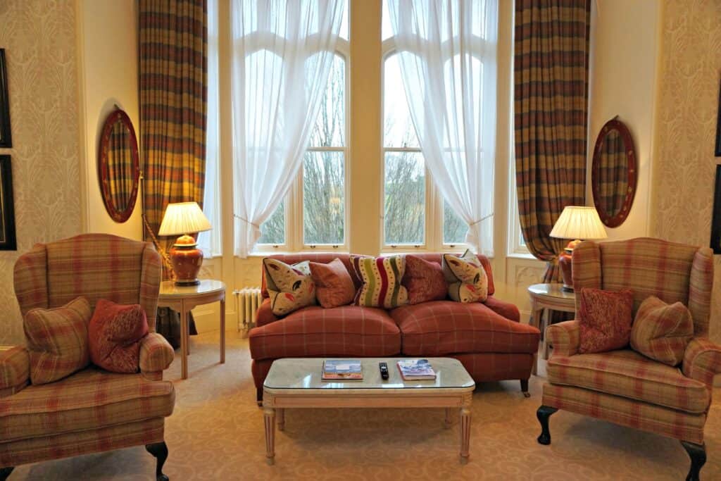 Dromoland Castle suite sitting area in front of window with plaid curtains - pink and cream colured love seat and high back chairs and glass top coffee table. Two tables with lamps behind.