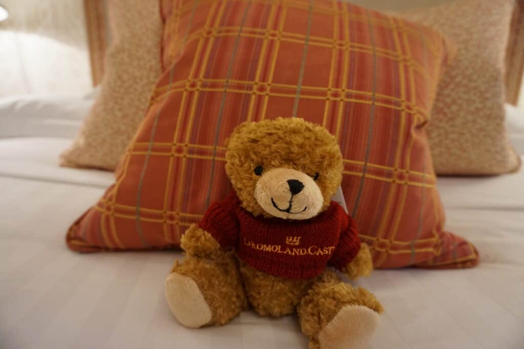 Teddy bear wearing red sweater with a crown and words Dromoland Castle sitting on white bed propped against pink plaid cushion.