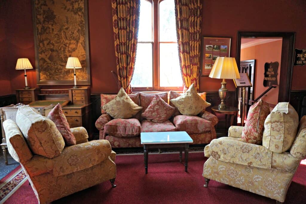 Drawing room at Dromoland Castle furnished with dark pink area rug, pink flowered sofa and cushions, two cream-coloured flowered chairs with pink cushions and small table. Wooden furniture and lamps at perimeter and window with curtains behind sofa.